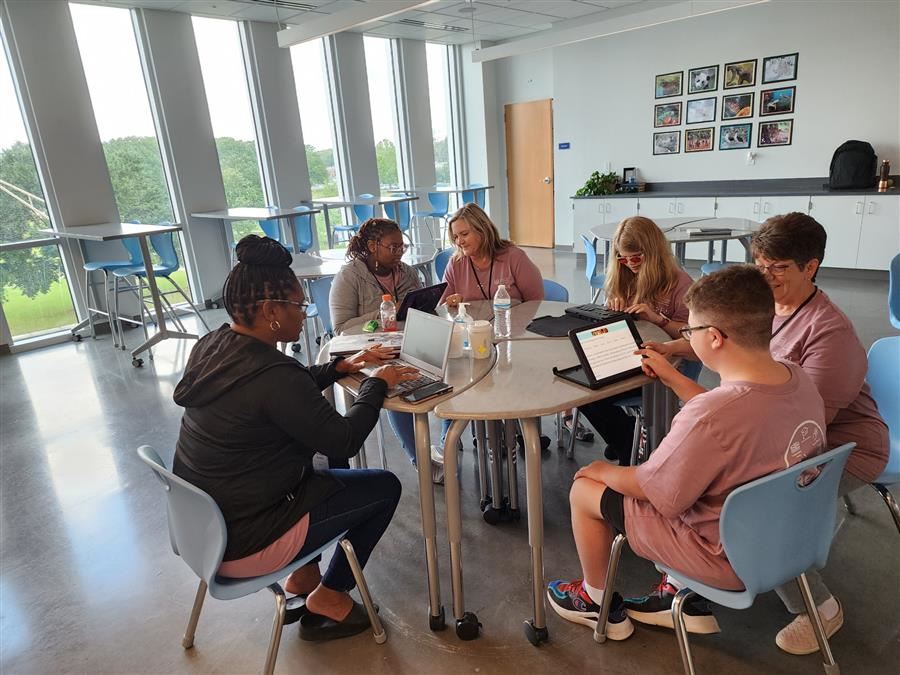 Middle school aged students and teachers sitting at a round table working on different AT devices. 